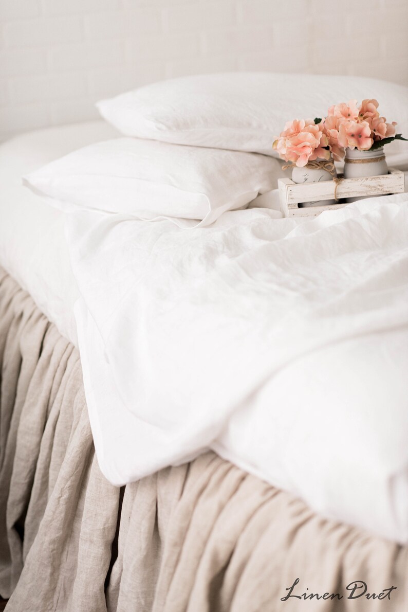 May include: White linen bedding with a tan linen bed skirt.  A small wooden tray with pink flowers in glass jars sits on the bed.