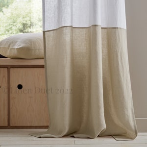May include: A pair of linen curtains with a two-toned design. The top half of the curtains is white, while the bottom half is a light beige color. The curtains are hanging in a window and are partially drawn.