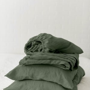 May include: A stack of sage green linen bedding, including a fitted sheet, flat sheet, and pillowcases. The bedding is folded neatly and arranged on a white surface. The text "Linen Duet" is visible in the bottom right corner of the image.