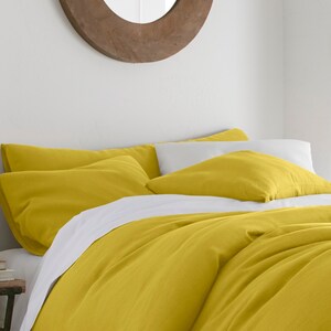 Puede incluir: Una funda n&oacute;rdica y fundas de almohada de lino amarillo en una cama con una s&aacute;bana blanca. La cama est&aacute; en un dormitorio con una pared blanca y un espejo de madera.