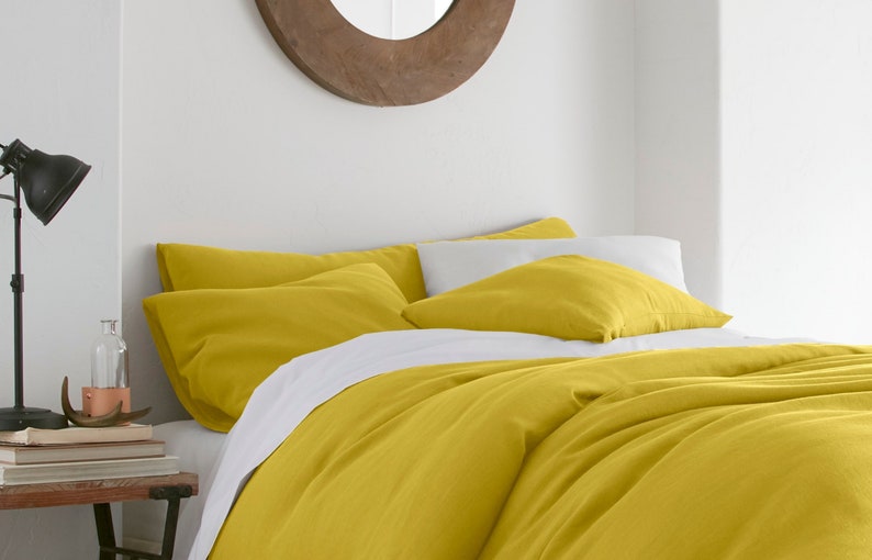 Puede incluir: Una funda n&oacute;rdica y fundas de almohada de lino amarillo en una cama con una s&aacute;bana blanca. La cama est&aacute; en un dormitorio con una pared blanca y un espejo de madera.