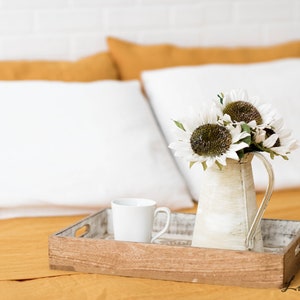 Puede incluir: Una bandeja de madera con una taza de cer&aacute;mica blanca y una jarra blanca llena de girasoles blancos sobre una colcha amarilla mostaza.