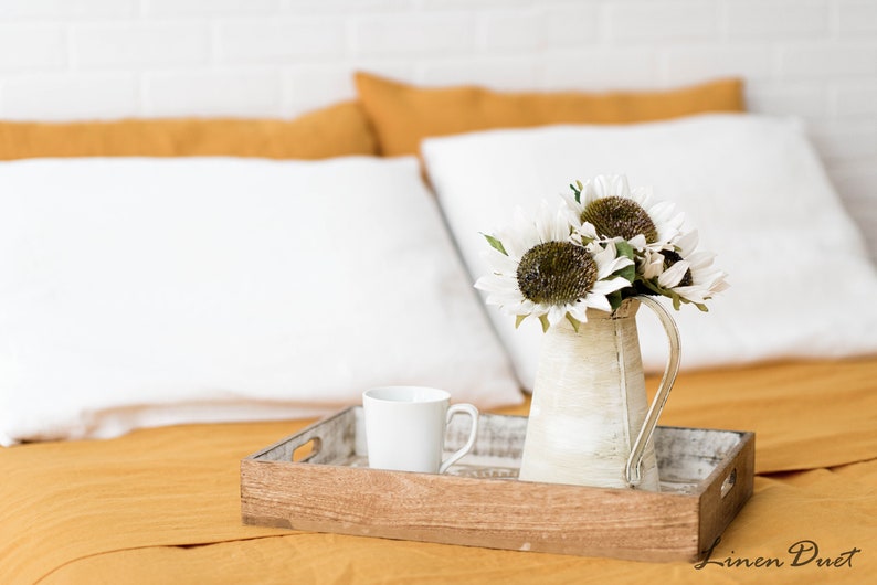 Puede incluir: Una bandeja de madera con una taza de cer&aacute;mica blanca y una jarra blanca llena de girasoles blancos sobre una colcha amarilla mostaza.