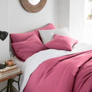 Puede incluir: Una funda n&oacute;rdica de lino rosa con fundas de almohada blancas y una s&aacute;bana blanca. La ropa de cama est&aacute; en una cama con una cabecera de madera y un espejo redondo en la pared detr&aacute;s de la cama.