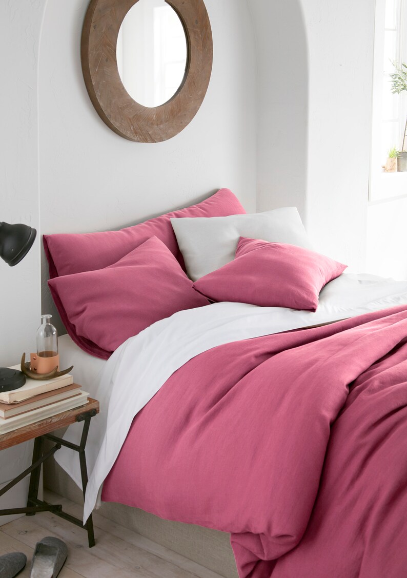 Puede incluir: Una funda n&oacute;rdica de lino rosa con fundas de almohada blancas y una s&aacute;bana blanca. La ropa de cama est&aacute; en una cama con una cabecera de madera y un espejo redondo en la pared detr&aacute;s de la cama.