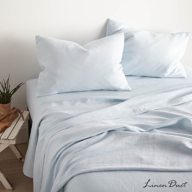 May include: A light blue linen sheet set with two matching pillowcases. The sheet is folded and the pillowcases are placed on top of the sheet. The sheet set is on a bed with a white headboard.