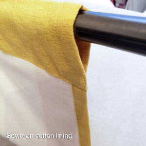 May include: Close-up of a yellow cotton lining sewn onto a white fabric panel. The fabric is hanging from a metal rod.