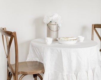 May include: A white tablecloth with a ruffled edge covers a round wooden table. Two white ceramic mugs and a white pie plate with a pie are on the table. A white vase with white flowers sits on the table. A wooden chair with a cross-back design is in the foreground.