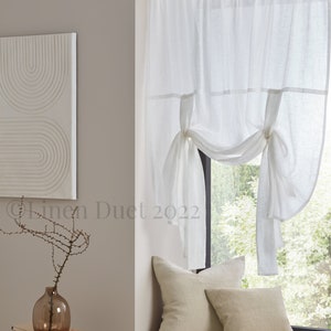 May include: White linen curtains with a gathered design, tied back on each side. The curtains are hung in a window with a view of trees and greenery. The window is in a room with a light grey wall and a wooden windowsill.