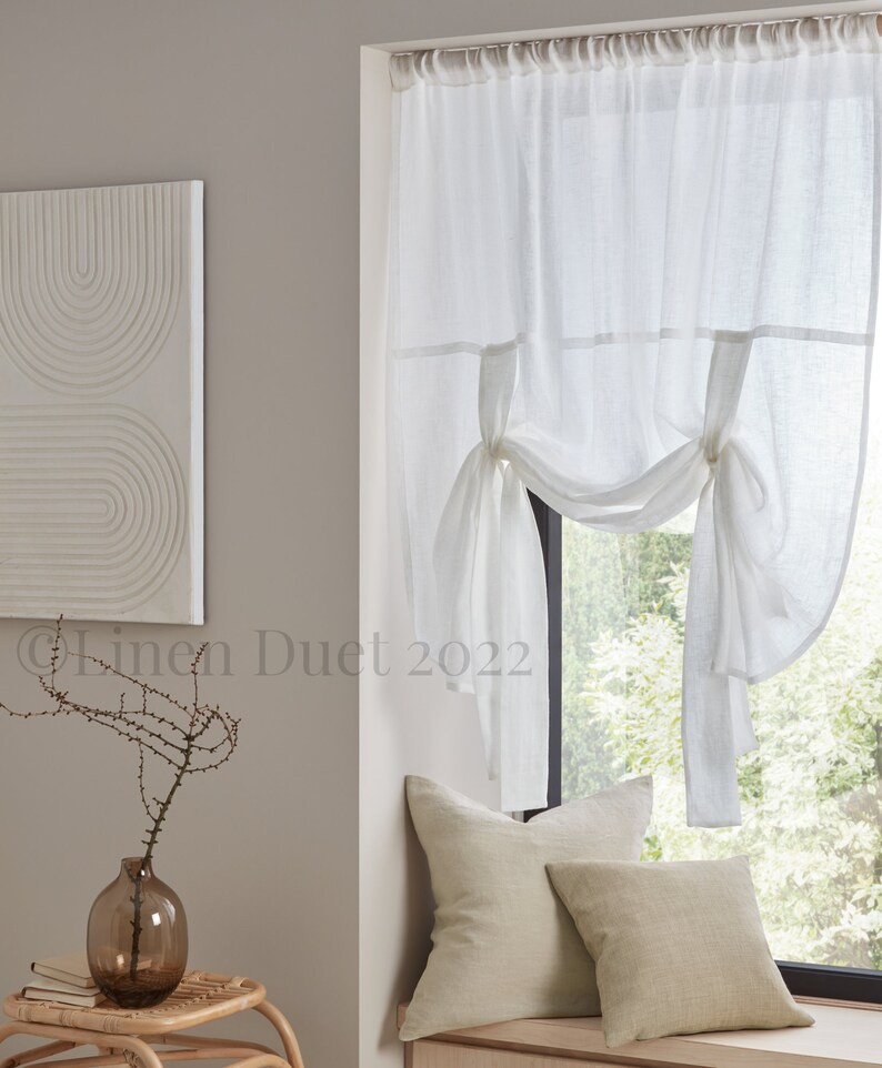 May include: White linen curtains with a gathered design, tied back on each side. The curtains are hung in a window with a view of trees and greenery. The window is in a room with a light grey wall and a wooden windowsill.