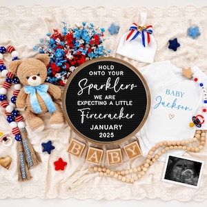 May include: A black letter board with the text "Hold onto your sparklers, we are expecting a little firecracker, January 2025".  Surrounding the letter board are various baby items, including a white onesie with the text "Baby Jackson" in blue, a blue and white baby hat, a wooden bead garland, a red, white, and blue beaded garland, a teddy bear, and a sonogram image.