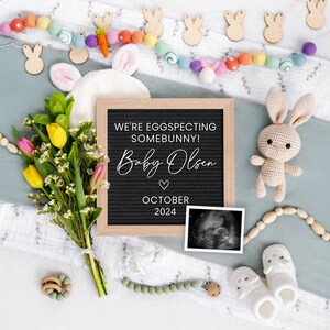 K&ouml;nnte beinhalten: Eine Buchstabentafel mit dem Text "We're eggspecting somebunny! Baby Olsen October 2024" umgeben von einem Strau&szlig; gelber und rosa Blumen, einem geh&auml;kelten Hasen und einem Baby-Ultraschallbild.
