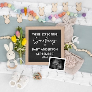 May include: A black letter board with the text "We're expecting somebunny" and "Baby Anderson September 2024". The board is surrounded by a variety of baby items, including a knitted hat, a wooden bunny rattle, a wooden bead garland, and a bouquet of flowers.