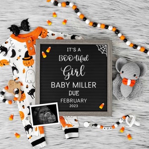 Puede incluir: Un tablero de letras negro con el texto "It's a BOO-tiful Girl Baby Miller Due February 2023".  Un murci&eacute;lago de crochet gris con una pajarita naranja est&aacute; en el lado derecho del tablero.  Un mameluco de beb&eacute; blanco y naranja con un estampado de Halloween negro y naranja est&aacute; en el lado izquierdo del tablero.  Hay ma&iacute;z dulce naranja y blanco y un collar de cuentas blanco y negro con un sonajero de fantasma de madera en el tablero.