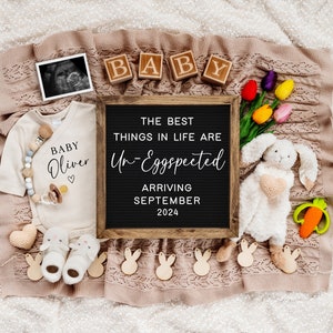 May include: A black letter board with the text "The best things in life are Un-Eggspected" and "Arriving September 2024".  The letter board is surrounded by a variety of baby-themed items, including a white onesie with the text "Baby Oliver", a stuffed bunny rabbit, and a string of wooden bunny shapes.