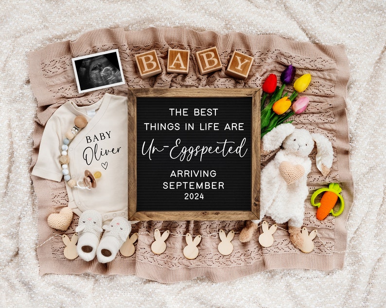 May include: A black letter board with the text "The best things in life are Un-Eggspected" and "Arriving September 2024".  The letter board is surrounded by a variety of baby-themed items, including a white onesie with the text "Baby Oliver", a stuffed bunny rabbit, and a string of wooden bunny shapes.