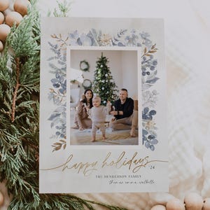 May include: A Christmas card with a watercolor border of blue and gold leaves. The card features a photo of a family of three standing in front of a Christmas tree. The text on the card reads "Happy Holidays 24" and "The Henderson Family Thomas, Emma, Isabella".