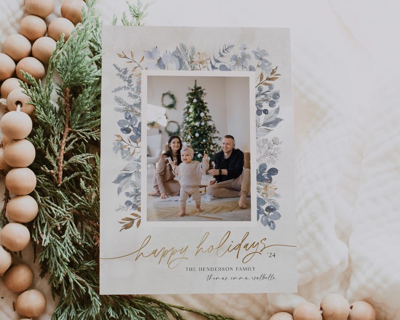May include: A Christmas card with a watercolor border of blue and gold leaves. The card features a photo of a family of three standing in front of a Christmas tree. The text on the card reads "Happy Holidays 24" and "The Henderson Family Thomas, Emma, Isabella".