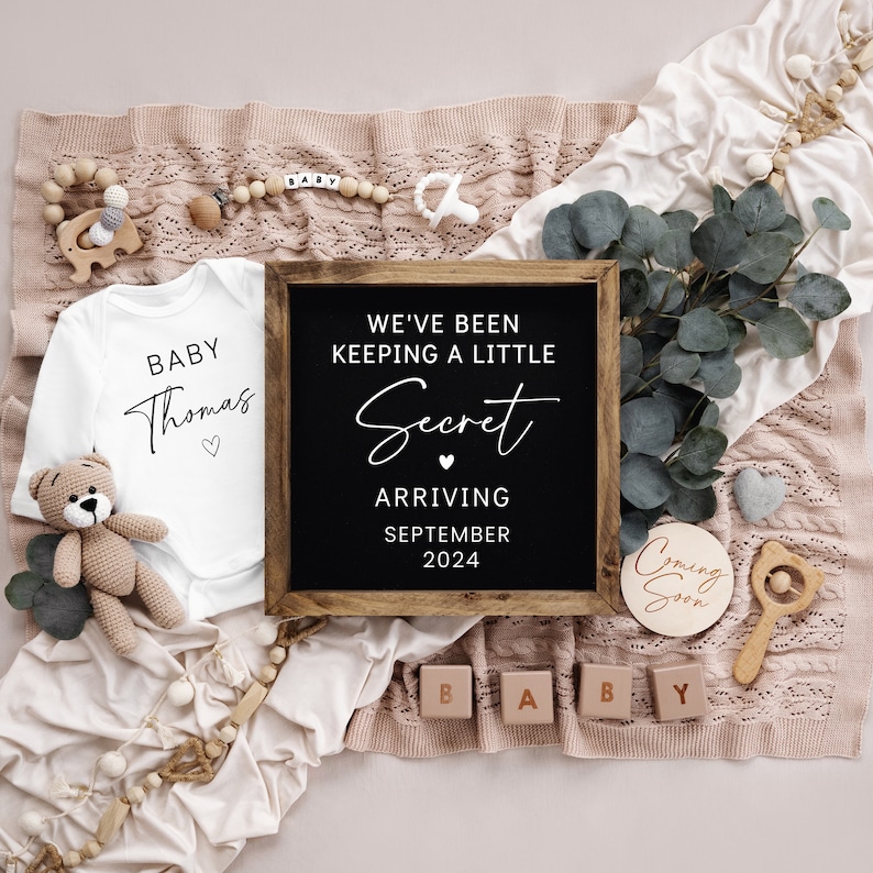 Op de afbeelding: Een baby-aankondiging met een wit rompertje met de tekst "Baby Thomas" en een zwart krijtbord met de tekst "We hebben een klein geheim bewaard. Aankomst in september 2024".