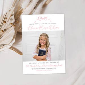 May include: A white invitation with a pink bow at the top. The text reads "Please join us for the baptism of Clara Rose Olsen." The invitation also includes a photo of a young girl holding a book. The text below the photo reads "Saturday, August 1, 2025 at 10:00 AM The Church of Jesus Christ of Latter-day Saints 125 North 100 West | Farmington"