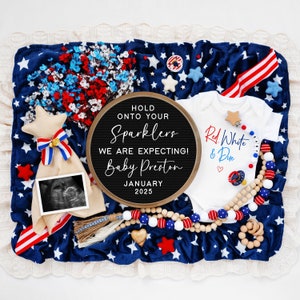 Op de afbeelding: Een zwart letterbord met de tekst "Hold onto your sparklers, we are expecting! Baby Preston, January 2025". Een wit rompertje met de tekst "Red, White & Due" in rood en blauw. Een kralenketting van hout met rode, witte en blauwe kralen. Een rammelaar voor baby's met een ster en het woord "Baby".