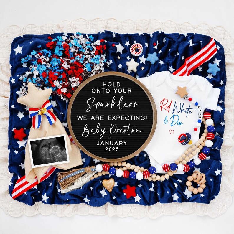 Op de afbeelding: Een zwart letterbord met de tekst "Hold onto your sparklers, we are expecting! Baby Preston, January 2025". Een wit rompertje met de tekst "Red, White & Due" in rood en blauw. Een kralenketting van hout met rode, witte en blauwe kralen. Een rammelaar voor baby's met een ster en het woord "Baby".