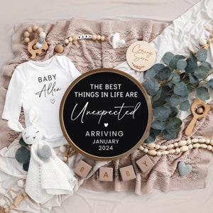 May include: A black and white sign with the text "The best things in life are unexpected" and "Arriving January 2024". A white onesie with the text "Baby Allen" is also in the image.