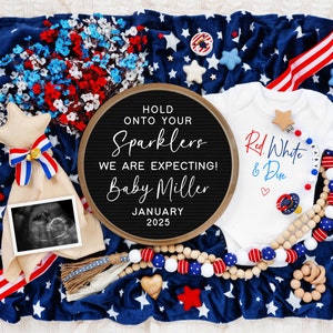 Op de afbeelding: Een rood, wit en blauwe baby romper met de tekst "Red, White & Due" en een hartje. Een letterbord met de tekst "Hold onto your sparklers, we are expecting! Baby Miller, January 2025". Een blauw en wit deken met sterren en een rood, wit en blauw lint. Een houten kralenketting met rode, witte en blauwe kralen. Een houten ster en een houten rammelaar.