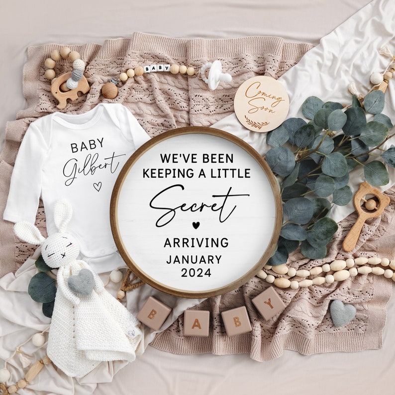 May include: A white onesie with the text "BABY Gilbert" and a heart. A wooden round sign with the text "WE'VE BEEN KEEPING A LITTLE Secret ARRIVING JANUARY 2024". The sign is surrounded by wooden blocks that spell out "BABY", a wooden elephant, a wooden rattle, a pacifier, and a garland of wooden beads.
