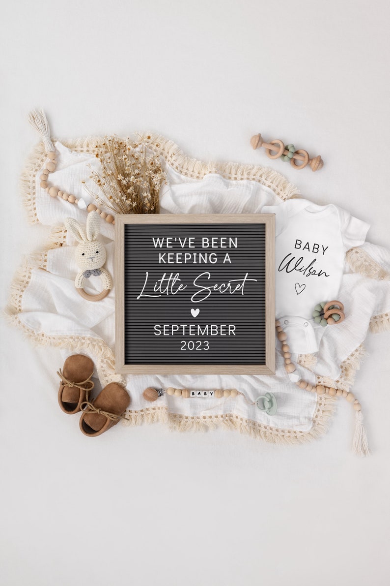 May include: A black letter board with the text "We've been keeping a little secret September 2023".  A white onesie with the text "Baby Wilson" is next to the letter board.  There are brown baby shoes, a wooden baby rattle, and a wooden bead garland with the word "baby" spelled out on it.