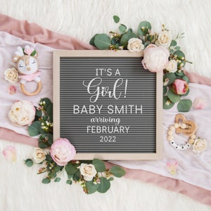 Może przedstawiać: Szara tablica z literami z tekstem "It's a Girl! Baby Smith arriving February 2022". Tablica jest otoczona r&oacute;żowymi i białymi kwiatami oraz zielenią. Jest drewniana grzechotka dla dziecka w kształcie jelenia i drewniany gryzak z koralikami.