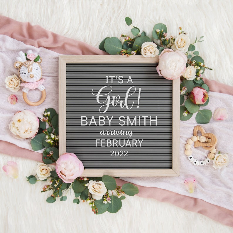 Może przedstawiać: Szara tablica z literami z tekstem "It's a Girl! Baby Smith arriving February 2022". Tablica jest otoczona r&oacute;żowymi i białymi kwiatami oraz zielenią. Jest drewniana grzechotka dla dziecka w kształcie jelenia i drewniany gryzak z koralikami.