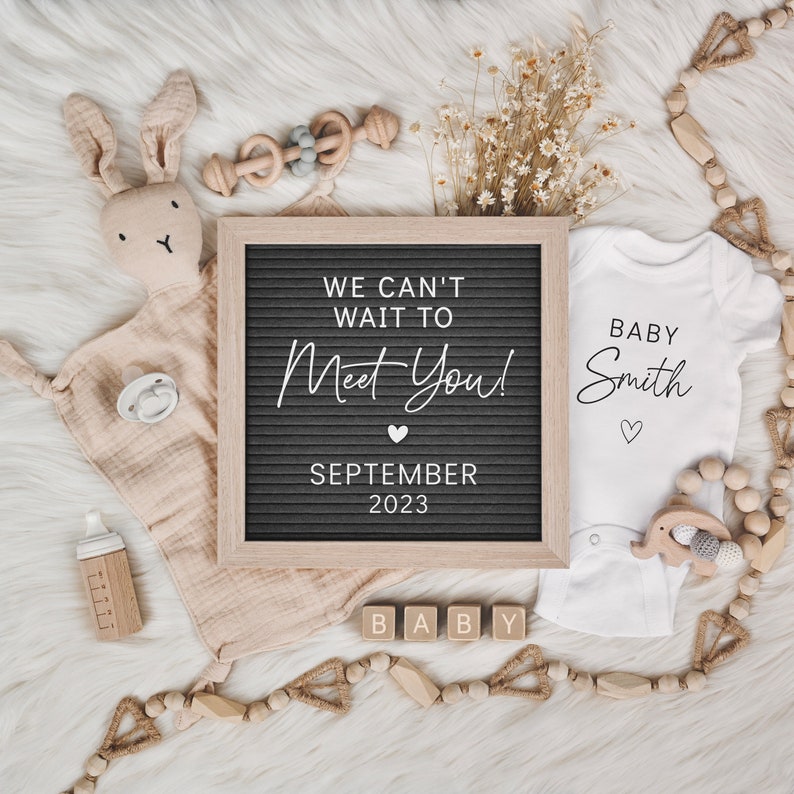 May include: A flat lay featuring baby announcement items. A letter board reads "WE CAN'T WAIT TO MEET YOU! SEPTEMBER 2023." A white onesie says "BABY Smith." Other items include a plush bunny, pacifier, and wooden blocks.