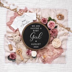 May include: A pink and white blanket with a white onesie that says "Baby Amelia" on it. There is a wooden sign that says "We are having a baby girl August 2024" and a small wooden sign that says "Baby Girl".