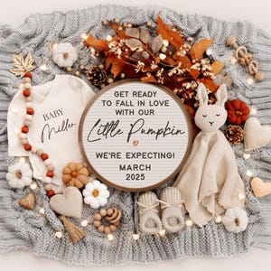 K&ouml;nnte beinhalten: Ein h&ouml;lzerner Buchstabenbrett mit dem Text "Get ready to fall in love with our little pumpkin. We're expecting! March 2025". Das Brett ist von Herbstdekorationen umgeben, darunter ein Baby-Strampler, eine Strickdecke und ein Stofftier-Kaninchen.