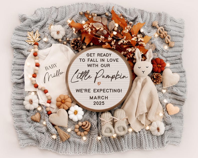 K&ouml;nnte beinhalten: Ein h&ouml;lzerner Buchstabenbrett mit dem Text "Get ready to fall in love with our little pumpkin. We're expecting! March 2025". Das Brett ist von Herbstdekorationen umgeben, darunter ein Baby-Strampler, eine Strickdecke und ein Stofftier-Kaninchen.