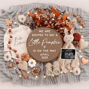 May include: A brown wooden sign with the text "We are excited to say a little pumpkin is on the way April 2025" surrounded by autumn-themed decorations, including a baby onesie, a stuffed bunny, and a pair of booties.