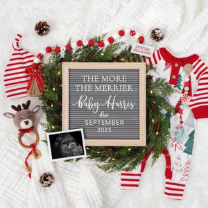 May include: A festive pregnancy announcement featuring a letter board with the text "The More The Merrier Baby Harris due September 2023". The announcement includes a baby onesie, a knitted reindeer toy, and a sonogram image, all arranged with Christmas decorations.