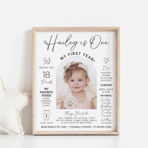 May include: A white framed photo with a black and white photo of a baby girl with a floral headband. The photo is surrounded by a white arch with text that says "Hailey is One" and "My First Year!"  The text also includes the baby's height and weight, favorite foods, and first milestones.