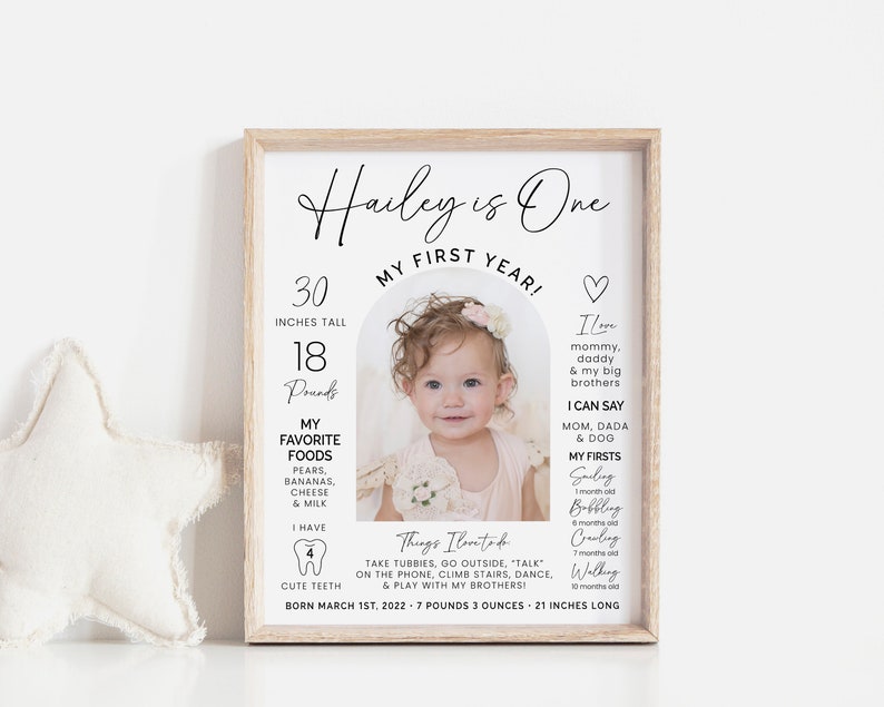 May include: A white framed photo with a black and white photo of a baby girl with a floral headband. The photo is surrounded by a white arch with text that says "Hailey is One" and "My First Year!"  The text also includes the baby's height and weight, favorite foods, and first milestones.