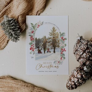 May include: A Christmas card with a watercolor wreath of greenery, berries, and white flowers surrounding a photo of a family of four in a snowy forest. The card reads "The Williams Family" and "Merry Christmas" with the names "Matthew, Lacy, Hazel, James" at the bottom.