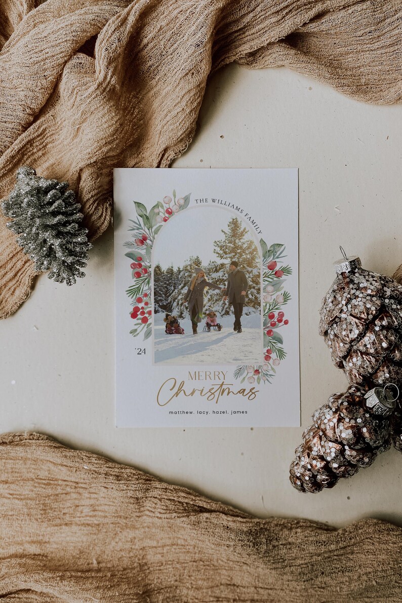 May include: A Christmas card with a watercolor wreath of greenery, berries, and white flowers surrounding a photo of a family of four in a snowy forest. The card reads "The Williams Family" and "Merry Christmas" with the names "Matthew, Lacy, Hazel, James" at the bottom.