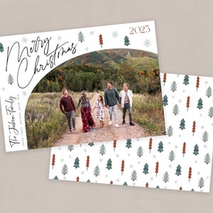 May include: A Christmas card featuring a family of five standing on a dirt road in front of a mountain range. The card has a white background with a green and red Christmas tree pattern and the text "Merry Christmas" and "The Jackson Family" in black cursive.