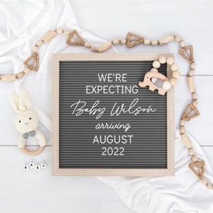 May include: A gray letter board with the text "We're expecting Baby Wilson arriving August 2022". A wooden elephant toy and a wooden bead garland are on the board. There are also wooden letter blocks that spell out "Baby" on a white surface.