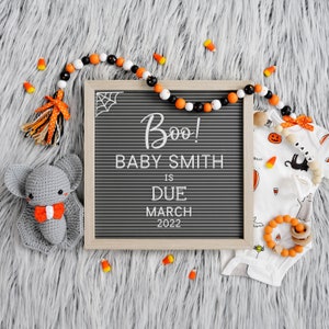 May include: A black letter board with the text "Boo! Baby Smith is due March 2022".  A gray crocheted bat, orange and black beads, and a white onesie with a black cat and pumpkin design are on a gray fuzzy surface.