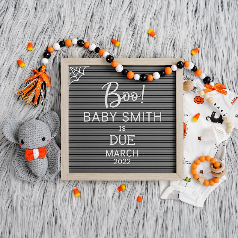 May include: A black letter board with the text "Boo! Baby Smith is due March 2022".  A gray crocheted bat, orange and black beads, and a white onesie with a black cat and pumpkin design are on a gray fuzzy surface.