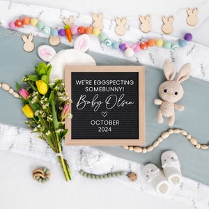 K&ouml;nnte beinhalten: Ein schwarzes Buchstabenbrett mit dem Text "We're eggspecting somebunny! Baby Olsen October 2024" umgeben von einer wei&szlig;en Decke, einem Strau&szlig; gelber und rosa Blumen, einem geh&auml;kelten Hasen und h&ouml;lzernen Hasendekorationen.