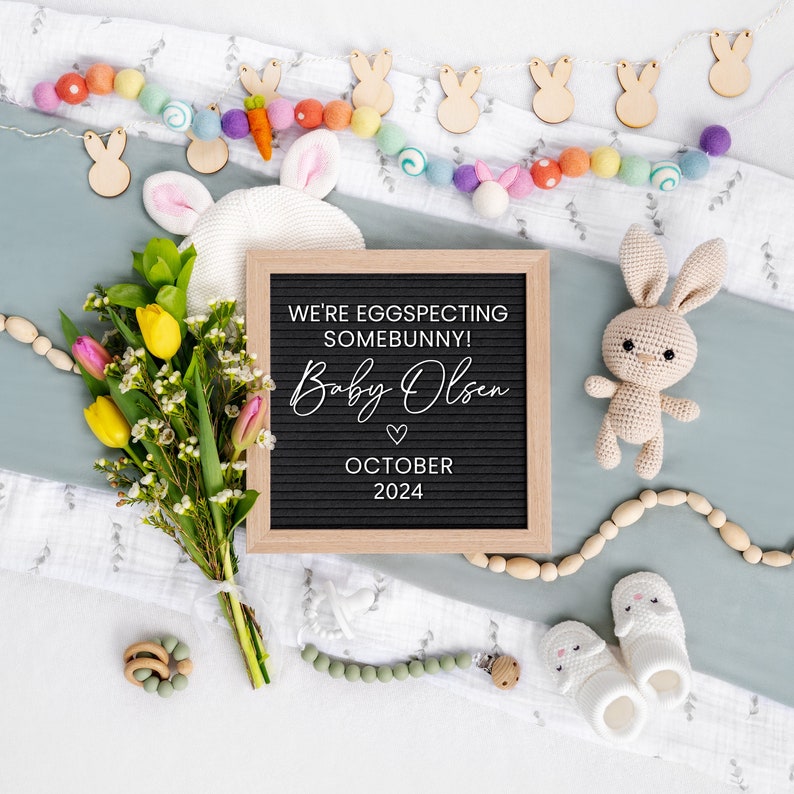 K&ouml;nnte beinhalten: Ein schwarzes Buchstabenbrett mit dem Text "We're eggspecting somebunny! Baby Olsen October 2024" umgeben von einer wei&szlig;en Decke, einem Strau&szlig; gelber und rosa Blumen, einem geh&auml;kelten Hasen und h&ouml;lzernen Hasendekorationen.