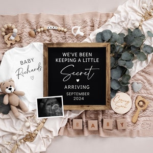 Op de afbeelding: Een baby-aankondiging met een wit rompertje met de tekst "Baby Richards" en een krijtbord met de tekst "We hebben een klein geheim bewaard, aankomst in september 2024". Er is ook een houten bord met de tekst "Binnenkort" en houten blokken die "Baby" spellen.