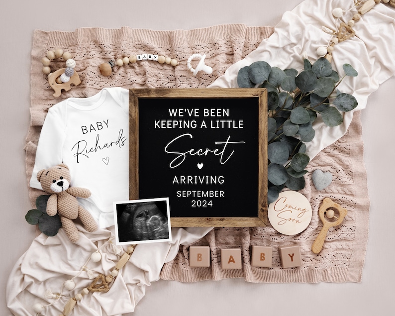 Op de afbeelding: Een baby-aankondiging met een wit rompertje met de tekst "Baby Richards" en een krijtbord met de tekst "We hebben een klein geheim bewaard, aankomst in september 2024". Er is ook een houten bord met de tekst "Binnenkort" en houten blokken die "Baby" spellen.